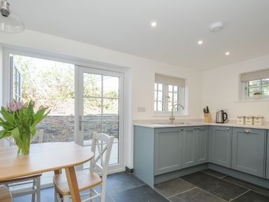 A kitchen with blue cabinets a wooden dining table with flowers and glass doors at Sunrise Cottage in St Kew Highway near Wadebridge