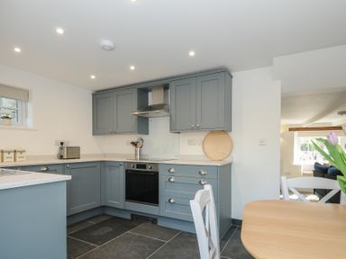 A kitchen with grey cabinets and black oven next to a dining table with white chairs and flowers at Sunrise Cottage in St Kew Highway near Wadebridge