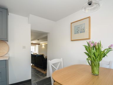 A dining area with wooden table and flowers in a vase near a white chair in kitchen view of living room beyond at Sunrise Cottage in St Kew Highway near Wadebridge
