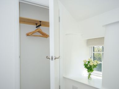 A small closet with wooden hangers and a window ledge with a vase of white roses at Sunrise Cottage St Kew Highway near Wadebridge