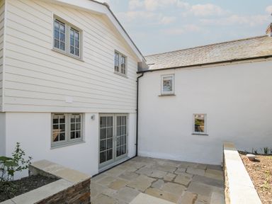 An outdoor patio area with stone paving and a white house with multiple windows and glass doors at Sunrise Cottage in St Kew Highway near Wadebridge