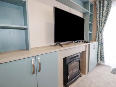 A TV on a wooden cabinet above an electric fireplace with built in shelves and blue cabinets near a window with patterned curtains at The Mermaid A04 in Par