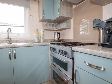 A kitchen with light blue cabinets, a stainless steel oven, a sink under a window, a kettle, stacked cups, glasses on shelves, and kitchen utensils at The Mermaid A04 in Par
