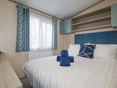 A bedroom with a bed with white bedding and blue pillows under cabinets and a window with blue patterned curtains at The Mermaid A04 in Par
