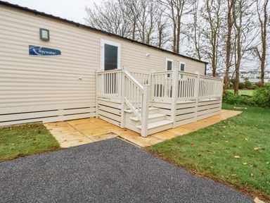 An exterior view of a beige mobile home with a small raised deck and stairs surrounded by grass and trees at The Mermaid A04 in Par