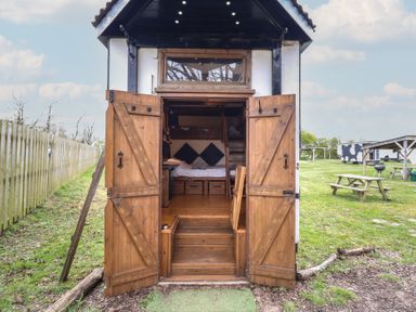 A small wooden cabin with open double doors showing a bed inside and picnic tables on grass outside at Daisy Cow Shed 1 in Easton near Campsea Ashe