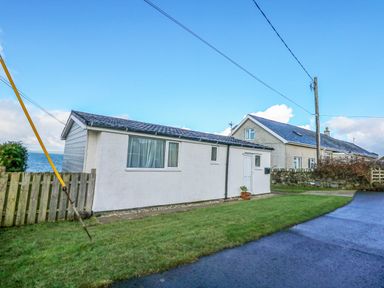A small white house with a grass lawn and a paved road nearby at Ael Y Don in Llandanwg