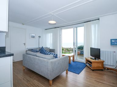 A living room with gray sofas blue cushions a small wooden TV stand with a flat screen TV a blue rug and sliding glass doors leading to a garden at Ael Y Don in Llandanwg