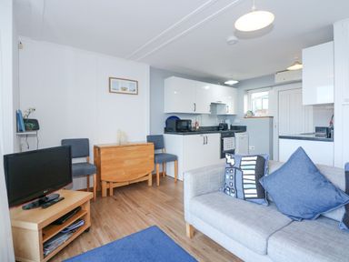 A living room with a gray sofa and two blue pillows a small wooden TV stand with a TV two chairs a foldable wooden table and a kitchen area with white cabinets at Ael Y Don in Llandanwg
