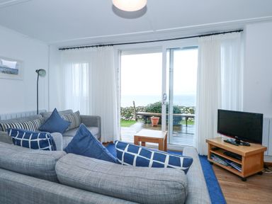 A living room with gray sofas and blue pillows a TV on a wooden stand and sliding glass doors with white curtains at Ael Y Don in Llandanwg