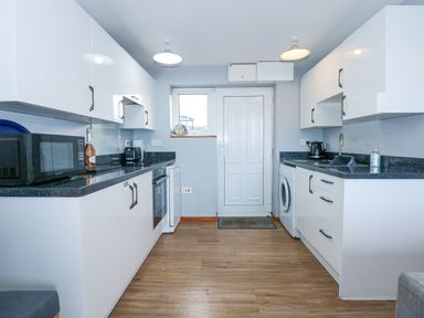A kitchen with white cabinets a microwave oven a bottle a toaster a stove a washing machine and a door at Ael Y Don in Llandanwg