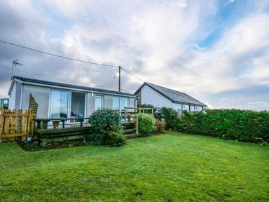 A backyard with grass lawn and bushes in front of a small house with sliding glass doors at Ael Y Don in Llandanwg