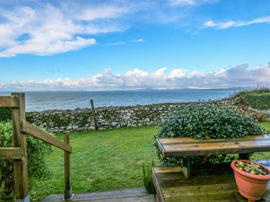 A garden with a wooden bench a potted plant grass and a stone wall overlooking the sea at Ael Y Don in Llandanwg