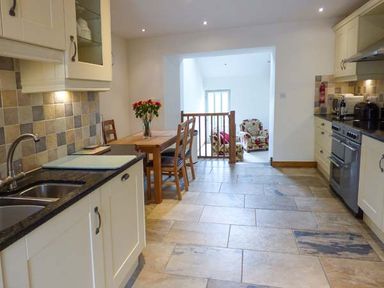 A kitchen with a tiled floor a wooden table with chairs and a floral armchair in the adjacent room at Ghyll Bank Barn in Staveley near Windermere
