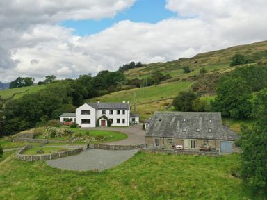 A white house and a stone barn in a rural hillside setting with green fields at Ghyll Bank Barn in Staveley near Windermere