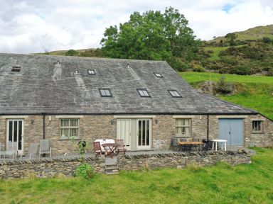 Stone building with slate roof chairs and tables on stone patio with grassy hill in background at Ghyll Bank Barn in Staveley near Windermere