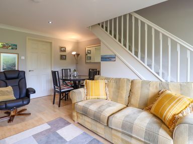 A living room with a striped sofa with yellow pillows a black armchair a round dining table with four chairs and a staircase at 50 Fernhill Heights in Charmouth