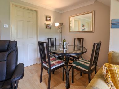 A dining area with a round glass table and four chairs with cushions a bottle and glasses on the table a large wall mirror and a floor lamp at 50 Fernhill Heights in Charmouth