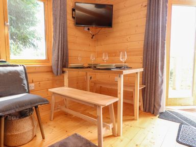 A wooden dining table with benches and plates set with wine glasses in a wooden room with a mounted TV and gray curtains at Northwood Lodge in Llanbedrog