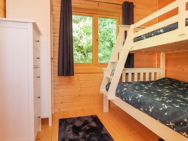 A bedroom with a white bunk bed a chest of drawers a wardrobe a window with dark curtains and a black rug on the wooden floor at Northwood Lodge in Llanbedrog