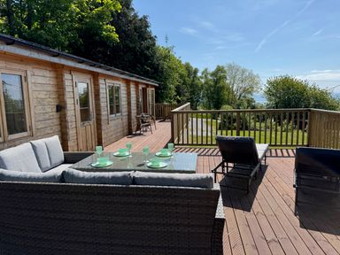 A wooden deck with outdoor seating and dining set next to a wooden cabin surrounded by trees at Northwood Lodge in Llanbedrog