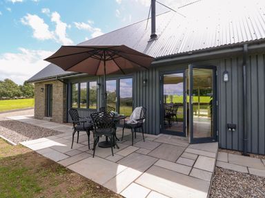 An outdoor patio with a table umbrella and chairs next to a building with glass doors at Strawberry Cottage Wester Essendy near Dunkeld