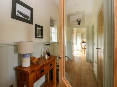 A hallway with a wooden console table holding a lamp and decor leading to a kitchen with cabinets and a dining table at Strawberry Cottage Wester Essendy near Dunkeld