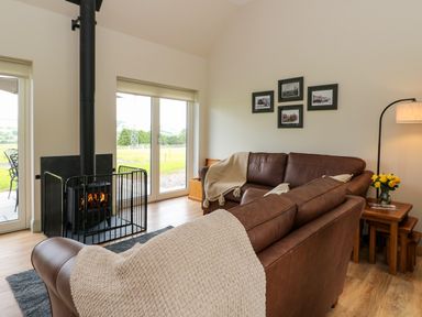 A living room with two brown sofas a wood burning stove behind a safety gate and a side table with a lamp and flowers at Strawberry Cottage Wester Essendy near Dunkeld