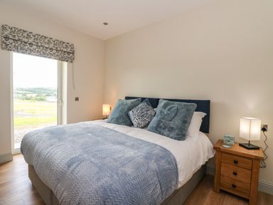 A bedroom with a large bed with blue cushions and blankets a wooden nightstand with a lamp and window with patterned curtain at Strawberry Cottage in Wester Essendy near Dunkeld