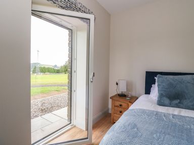 A bedroom with a bed blue bedding and a wooden nightstand next to an open glass door showing a green outdoor area at Strawberry Cottage Wester Essendy near Dunkeld