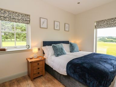 A bedroom with a double bed and bedside table with lamp near windows showing outdoor views at Strawberry Cottage in Wester Essendy near Dunkeld