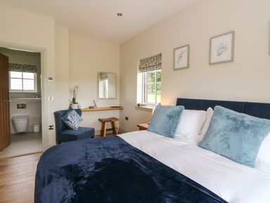 A bedroom with a bed with blue pillows and blanket a chair with a pillow a small wooden stool and a bathroom visible through an open door at Strawberry Cottage in Wester Essendy near Dunkeld