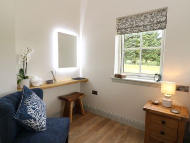 A small room with a blue chair and cushion a wooden stool a floating wooden shelf with decorative items a window with patterned blind and a wooden bedside table with a lamp at Strawberry Cottage in Wester Essendy near Dunkeld