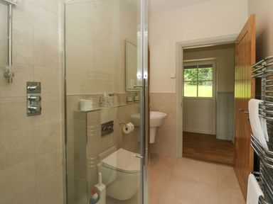 A bathroom with a shower, toilet, sink, open wooden door, and a window showing green grass at Strawberry Cottage in Wester Essendy near Dunkeld