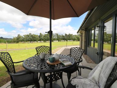 An outdoor patio with a round metal table and four chairs under an umbrella with a teapot cups and a book on the table at Strawberry Cottage Wester Essendy near Dunkeld