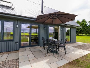 A patio with a round table and four chairs under an umbrella outside a building with grey walls and large glass doors at Strawberry Cottage Wester Essendy near Dunkeld