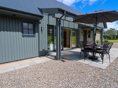 An outdoor patio area with a round table and four chairs under an umbrella next to a building with sliding glass doors at Bramble Cottage in Wester Essendy near Dunkeld