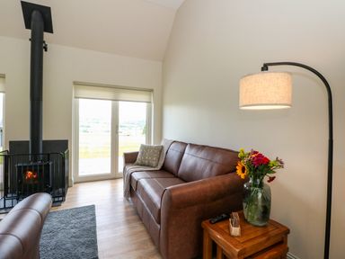 A living room with a brown leather sofa a wood stove glass doors a floor lamp and a side table with a vase of flowers at Bramble Cottage Wester Essendy near Dunkeld