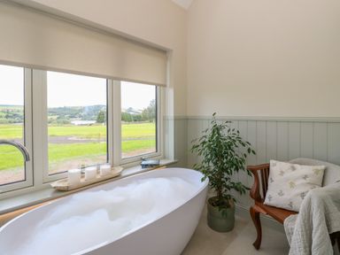 A bathroom with a bubble-filled bathtub near a window with candles on the sill and a wooden chair with a cushion and blanket at Bramble Cottage Wester Essendy near Dunkeld