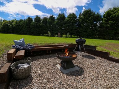 An outdoor seating area with a fire pit grill basket and wooden benches on gravel with trees in the background at Bramble Cottage in Wester Essendy near Dunkeld
