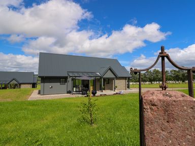 A modern house with a large sloped roof on a grassy lawn with a stone and metal structure in the foreground at Bramble Cottage Wester Essendy near Dunkeld