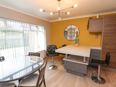 A dining area with a round glass table and chairs near large windows and a kitchen island with bar stools and a plant at 315 Filey Road in Scarborough