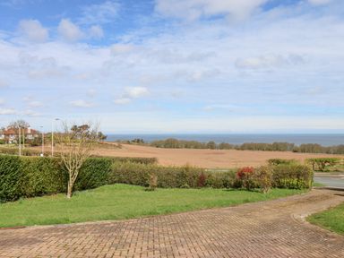A view of a brick driveway with grass and bushes leading to a road with fields and the sea in the background at 315 Filey Road in Scarborough