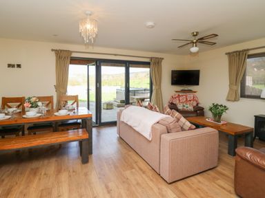 An open living room and dining area with a sofa, wooden dining table with benches, armchair, wall-mounted TV, and large sliding glass doors at Caban Dulas in Rhayader