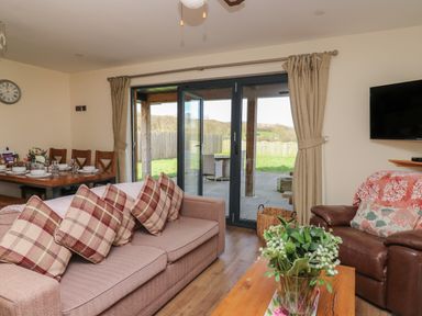 A living room with a brown sofa with plaid cushions a brown leather armchair a wall-mounted TV and dining table with chairs near glass doors at Caban Dulas in Rhayader