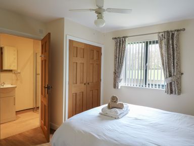 A bedroom with a bed and folded towels on it a window with curtains a ceiling fan wooden closet doors and an open door leading to a bathroom at Caban Dulas in Rhayader