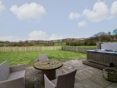 An outdoor patio with wicker chairs around a wooden cable spool table next to a hot tub and a grassy fenced garden at Caban Dulas in Rhayader