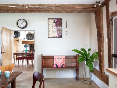 A room with a wooden bench with a pink flamingo cushion a plant in a white pot on a stand and a clock above an open doorway at Brook Farm in St Nicholas near Halesworth
