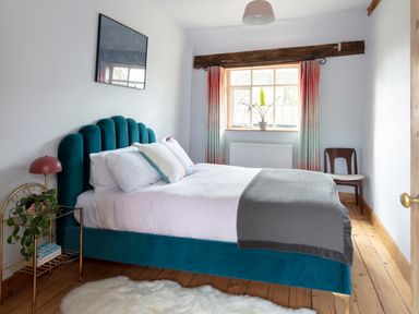 A bedroom with a teal upholstered bed white bedding a grey throw a wooden floor and a window with curtains at Brook Farm in St Nicholas near Halesworth