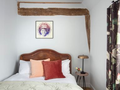 A bedroom with a wooden bed headboard red and peach pillows a small round side table with a lamp and flowers and a framed portrait on the wall at Brook Farm in St Nicholas near Halesworth